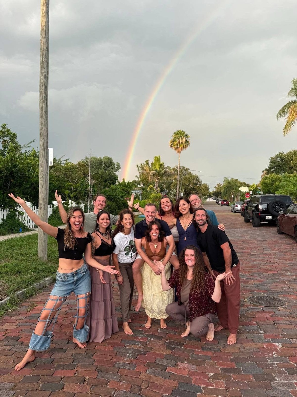 Megan and a joyful group of friends gathered barefoot on a brick street under a rainbow, arms outstretched.