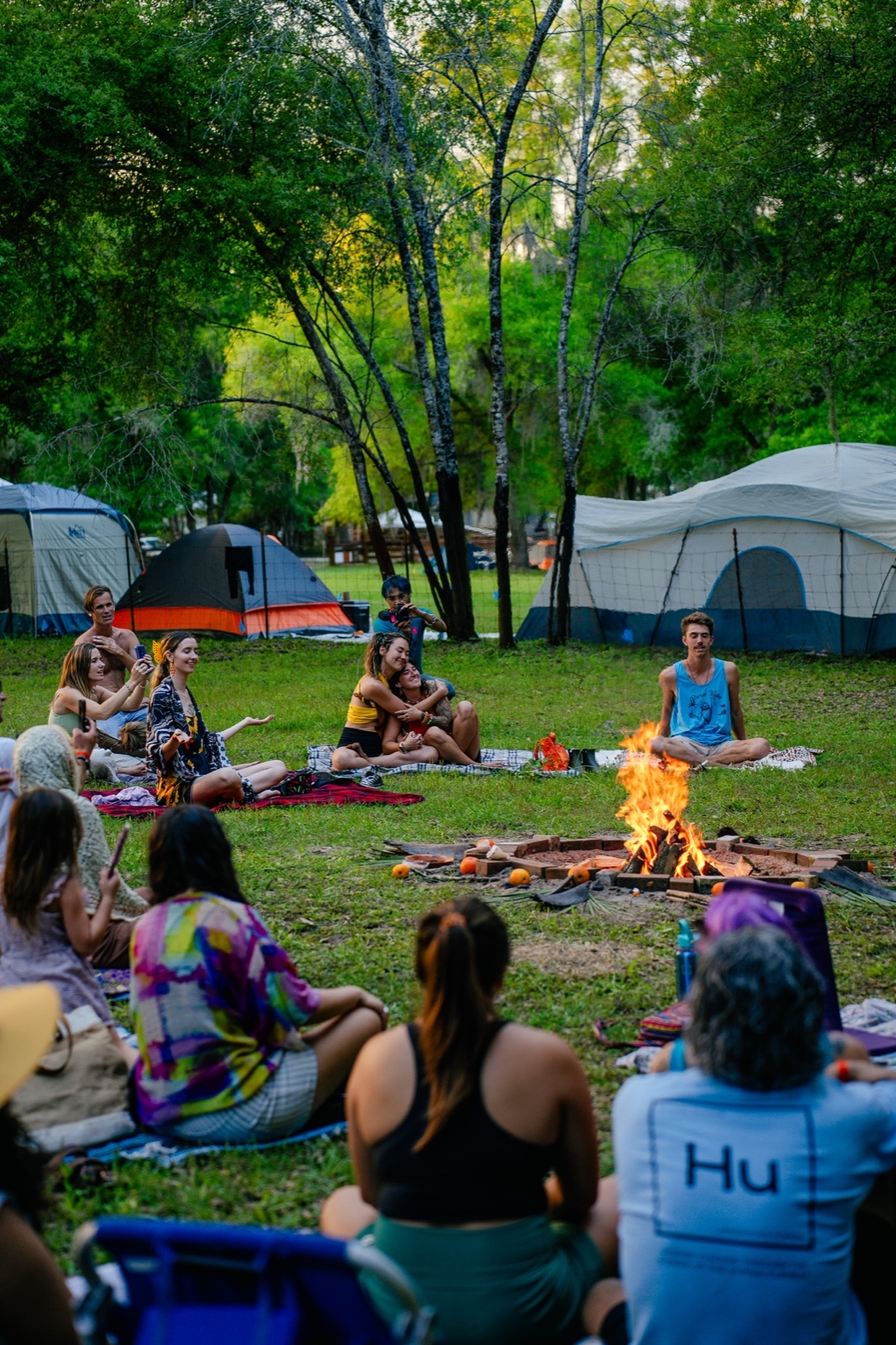 A group gathered around a campfire at a retreat, tents in the background among the trees.